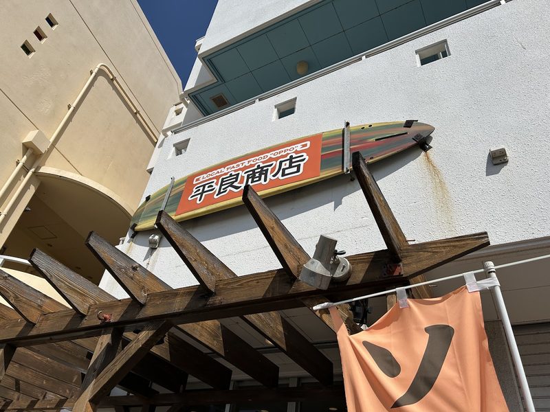 建物の横から見た木のテラスとサーフボード形の看板